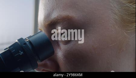 Close up of eyes of female doctor looking under microscope while conducting pharmacological test on samples in medical science laboratory. Development of technology and medicine. Health care concept. Stock Photo