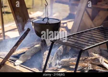 Black cast iron dutch oven hangs over a campfire cooking food at an outdoor campsite Stock Photo