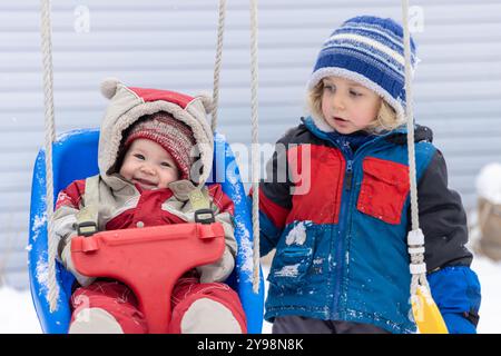 Toddler boy is pushing his baby sister on a swingset in the snow while wearing warm winter clothes. Selective focus Stock Photo
