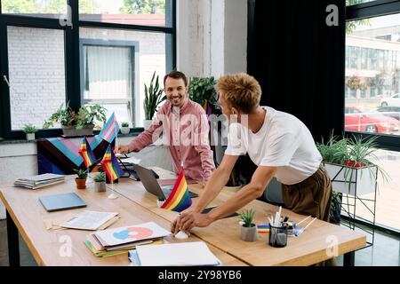 Two smiling men collaborate at a stylish desk adorned with pride flags and plants, sharing ideas. Stock Photo
