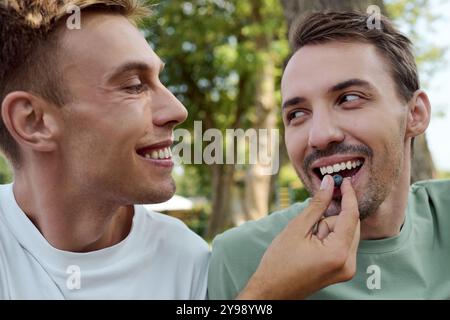 A happy couple shares a playful moment, smiling and feeding each other in a sunlit park. Stock Photo