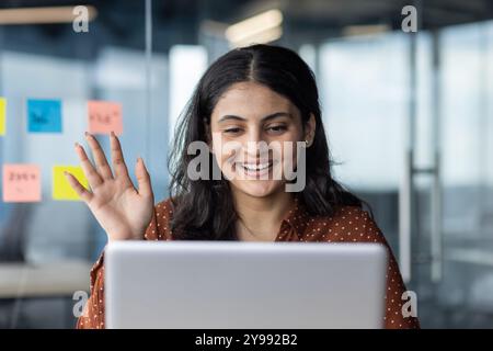 Smiling woman waves at laptop screen during virtual meeting, showcasing positive communication in modern office. Sticky notes on glass wall complement her cheerful engagement in digital workspace. Stock Photo
