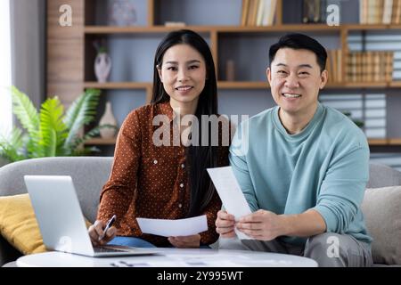 Asian couple sitting on sofa with documents. Smiling and discussing finances, using laptop for planning. Relaxed atmosphere at home. Stock Photo