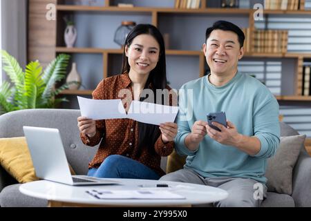 Cheerful asian couple trusts each other reviewing financial documents with phone and laptop. Together they plan budget with teamwork, happiness, and technology in home setting. Stock Photo