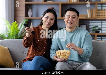 Asian couple enjoying movie night, sitting on couch, watching television, eating popcorn, suggesting leisure time. Highlights happiness, comfort, togetherness, family in living room setting. Stock Photo