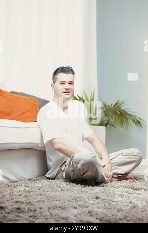 Man is sitting on the floor in a cross legged position, expressing positive emotions and enjoying the comfort of his home Stock Photo