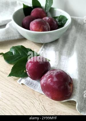 Fresh ripe plums in a ceramic bowl with leaves on a light wooden surface. Image for menu and cafe design Stock Photo