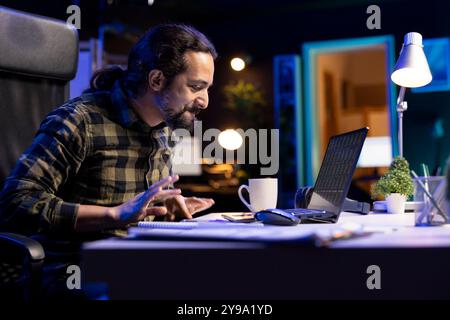 In his comfortable apartment, bearded man diligently works from home on his laptop, focused on financial research. Male freelancer carefully analyzes investment charts and studies market trends Stock Photo