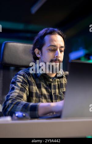 Serious millennial man using laptop sitting at the table, focused casual guy communicating online, writing emails, distantly working or studying on his portable computer in his apartment. Stock Photo