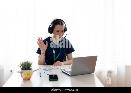 Happy young general practitioner in headphones using laptop, chatting to patient on video call, giving help, online consultation, advice, attending vi Stock Photo