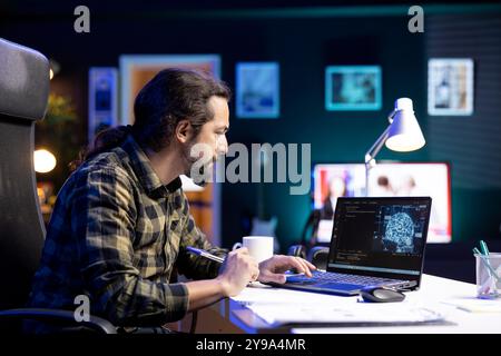 Male programmer coding for AI and machine learning on digital laptop. Bearded man looking at neural network and database on his electronic device while comparing with information on his notebook. Stock Photo