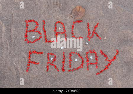 Black Friday words made with small red stones, on a sand background. Small shells and a dried leaf also appear in the scene. Stock Photo