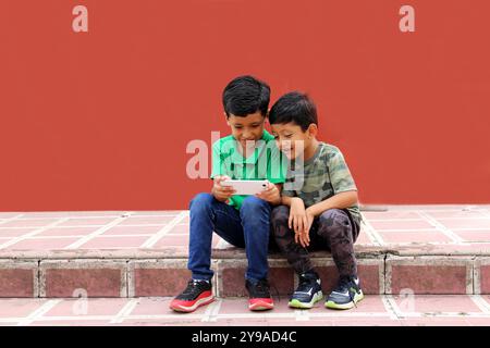 Two dark-haired Latino children who are brothers or friends who live in poverty play video games with a cell phone, make video calls and take selfies Stock Photo