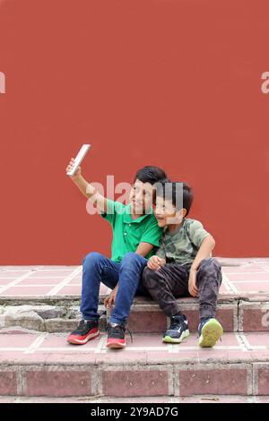 Two dark-haired Latino children who are brothers or friends who live in poverty play video games with a cell phone, make video calls and take selfies Stock Photo