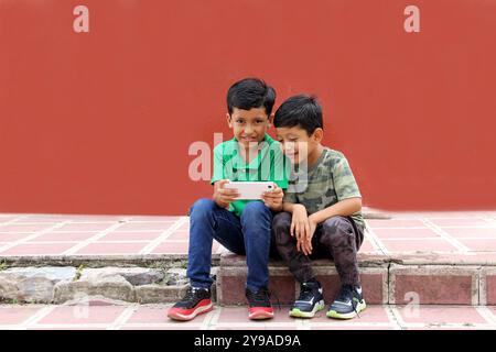 Two dark-haired Latino children who are brothers or friends who live in poverty play video games with a cell phone, make video calls and take selfies Stock Photo