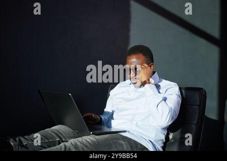 Portrait Of Pensive African Business Man In Glasses Sitting At Desk With Laptop In Home Office, Looking Away With Glad Face Expression. Smiling Guy In Stock Photo