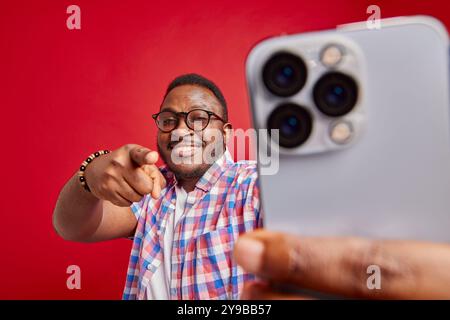 Young african american man pointing finger at phone while holding it in front of him on isolated red background, studio portrait Stock Photo