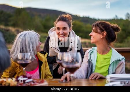 Group of diverse women friends laughing and enjoying wine tasting outdoors. Scenic mountain backdrop, casual attire, and warm social atmosphere captur Stock Photo
