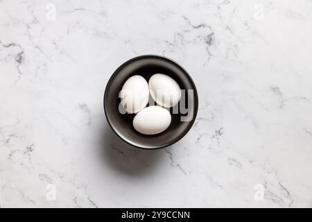 Top view of three white eggs in a vintage black bowl on a marble background. Stock Photo