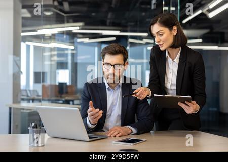 Colleagues in formal attire engaged in business meeting. Woman pointing at laptop screen, man contemplating solution. Office desk with laptop, smartphone, and documents enhancing collaborative Stock Photo