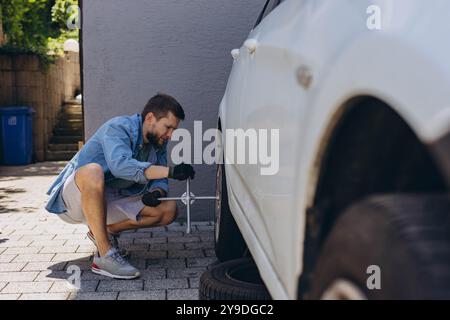 Male auto mechanic in unscrewing lug nuts on car wheel in process of new tire replacement, using wrench while changing flat tire on the road. High qua Stock Photo