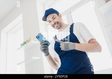 Smiling handyman in overalls using smartphone during home renovation project in bright modern room Stock Photo