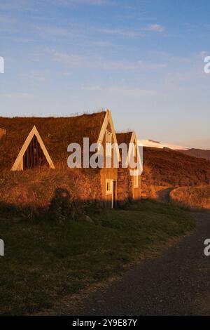 Charming turf houses bask in golden sunset light, nestled in a serene Icelandic landscape with a winding path leading to the countryside. Stock Photo