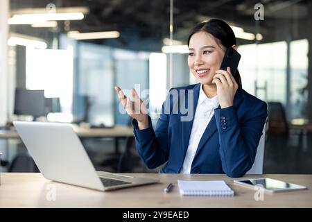 Asian business woman in office using phone while multitasking with laptop and taking notes, showcasing confidence, productivity, technology, communication, and professional work environment. Stock Photo