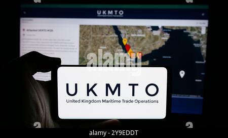 Person holding cellphone with logo of United Kingdom Maritime Trade Operations (UKMTO) on screen in front of webpage. Focus on phone display. Stock Photo