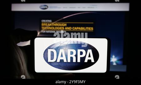 Person holding cellphone with logo of Defense Advanced Research Projects Agency (DARPA) on screen in front of webpage. Focus on phone display. Stock Photo