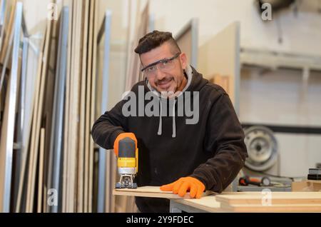 Carpenter man cutting wood in his workshop with a hand jigsaw, dressed in black work clothes, safety glasses and orange gloves Stock Photo