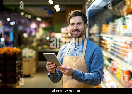Grocery store employee wearing apron holding tablet in supermarket aisle. Man smiling, using technology to manage inventory and enhance customer service. Stock Photo