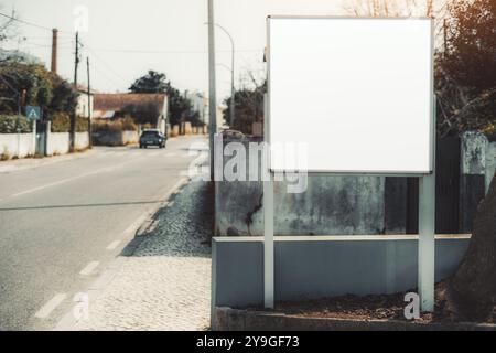 A suburban street with an empty billboard for mockup display. The road is lined with houses and trees, while a car drives in the distance. Blank billb Stock Photo