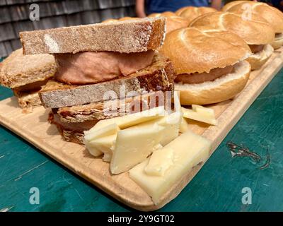A wooden cutting board on a green table with Leberkaese rolls and bread and pieces of cheese Stock Photo