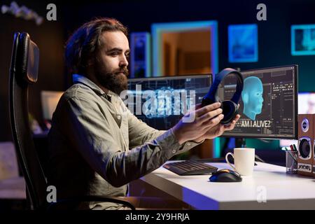 IT specialist putting headphones on to be able to listen to artificial intelligence speaking on computer. Programmer using audio device to communicate with awaken AI on PC asking existential questions Stock Photo