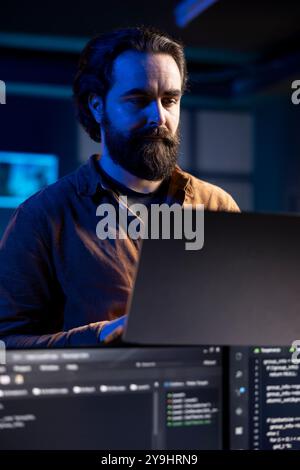 Teleworking IT admin working from RGB lit home using laptop, testing and deploying programs and systems. Software technician checking code displayed on computer screens, standing in apartment Stock Photo