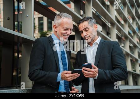 Mature businessmen in suits standing outdoors, using smartphones for work related tasks, sharing information and discussing business strategies in a modern urban setting. Professional collaboration and mobile technology. High quality photo Stock Photo