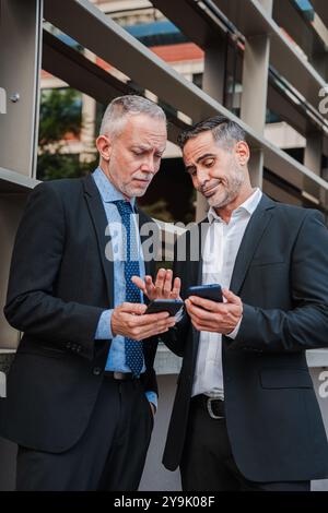 Vertical. Mature businessmen in suits standing outdoors, using smartphones for work related tasks sharing information, discussing business strategies. Professional collaboration and mobile technology. High quality photo Stock Photo