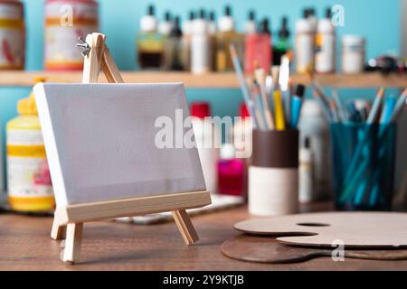Front view of small wooden easel with blank canvas on wooden table against background of shelves with paint tubes, brushes and pencils Stock Photo