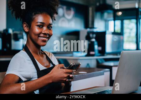 Close up portrait of concentrated black woman coffee shop owner in apron using smartphone in her Stock Photo