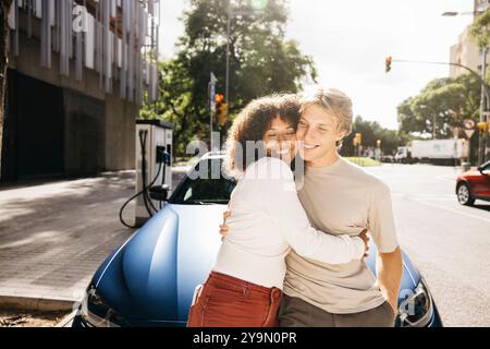 A joyful couple enjoys a moment together while their electric car charges in an urban setting. They are happy and relaxed, showcasing sustainable livi Stock Photo