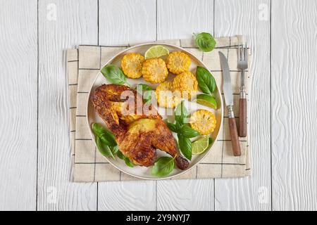 half of bbq chicken with corn cobs coins and fresh basil leaves on plate on white wooden table with cutlery, horizontal view from above, flat lay Stock Photo