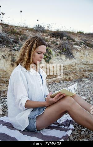 Beautiful woman reading a book sitting on a pebble beach vertical Stock Photo
