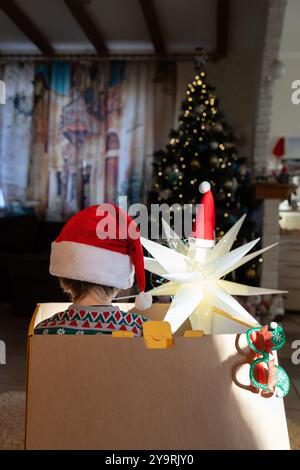 unrecognizable child in Santa hat sits in large cardboard box with glowing lantern in room in front of Christmas tree. waiting for Christmas miracle. Stock Photo