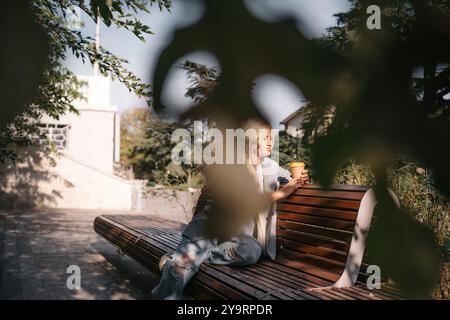 A blonde woman sits on a bench with a cup of coffee in her hand. She is wearing a gray jacket and jeans. The bench is wooden and has a curved back Stock Photo