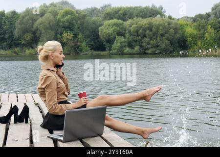 Woman office worker sitting on a pier by the river drinking coffee, talking on the phone and splashes water with his feet near the laptop. The concept Stock Photo