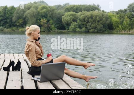 Woman office worker sitting on a pier by the river drinking coffee, talking on the phone and splashes water with his feet near the laptop. The concept Stock Photo