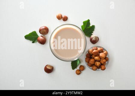 Glass of hazelnut drink isolated on white table with bowl of peeled and shelled nuts and leaves around. Top view. Stock Photo