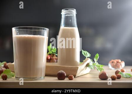Detail of hazelnut drink in a glass on wooden kitchen bench and full bottle on a tray and nuts and leaves around with dark kitchen in background. Fron Stock Photo