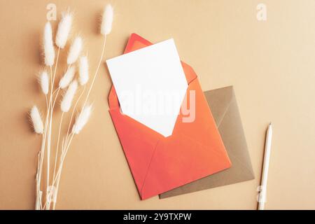 Red envelope with blank card, pencil and bunny tails grass Lagurus Ovatus flowers on brown background. Top view, flat lay, mockup. Stock Photo